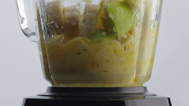 Ηλεκτρικό μπλέντερ προετοιμασία νόστιμο μείγμα βιταμινών φρούτων σε σούπερ αργή κίνηση από κοντά. Ζουμερά φρέσκα μπανάνα μήλο πορτοκαλί ανάμειξη σε γυάλινο μίξερ λευκό φόντο. Μαγειρική χορτοφάγος υγιεινά τρόφιμα. - Φωτογραφία, εικόνα