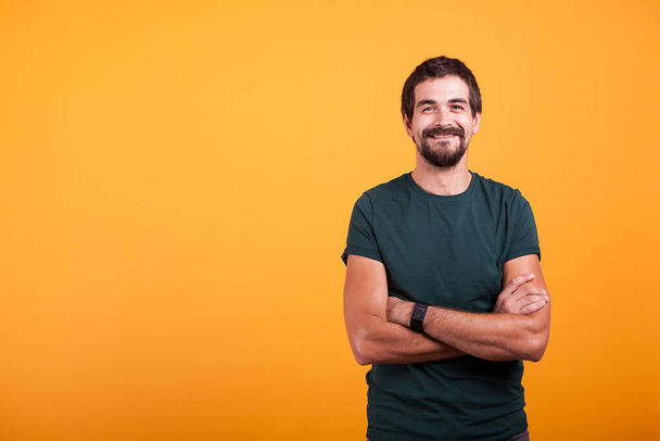 Handsome man with his arms crossed smiling at the camera isolated on yellow background. Portrait of attractive bearded confident person in studio photo - Foto, afbeelding