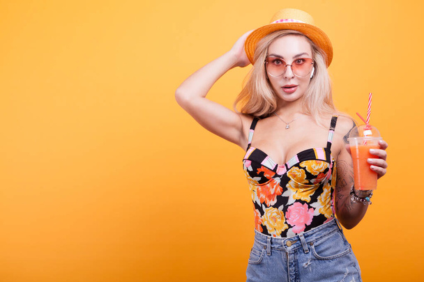 Attractive blonde haired woman showing orange juice with sunglasses over yellow background - Foto, immagini