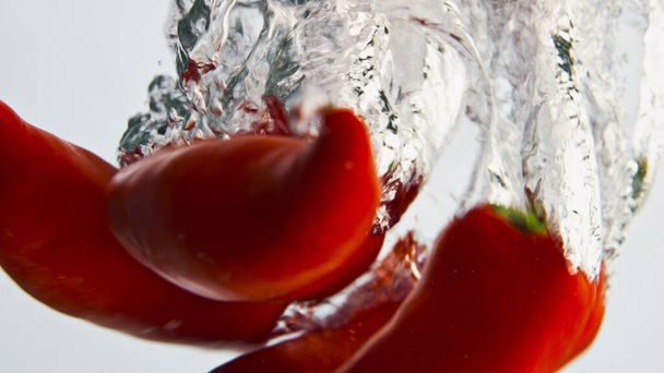 Red peppers splashing liquid closeup. Beautiful spicy vegetables fall bubbling water in light background. Hot chili bounce in container. Healthy raw eating. Beautiful organic food commercial shot. - Fotó, kép