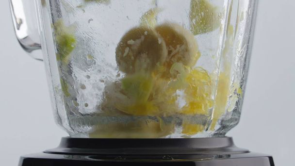 Ρίχνουμε κομμάτια φρέσκων νόστιμων φρούτων στο μπλέντερ με περιστρεφόμενες μεταλλικές λεπίδες από κοντά. Κομμένα μήλα πορτοκαλί μπανάνα ανάμειξη στο μπολ μίξερ γυαλί σούπερ αργή κίνηση. Προετοιμασία νόστιμη διατροφή βιταμινών. - Φωτογραφία, εικόνα