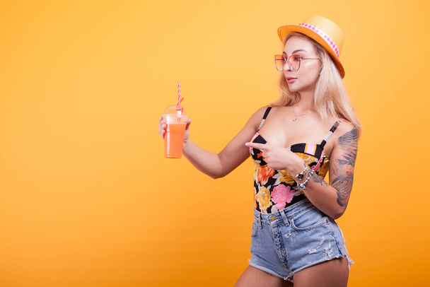 feliz jovem mulher olhando para o verão fresco suco de laranja pensando ela é sexy no estúdio sobre fundo amarelo
 - Foto, Imagem
