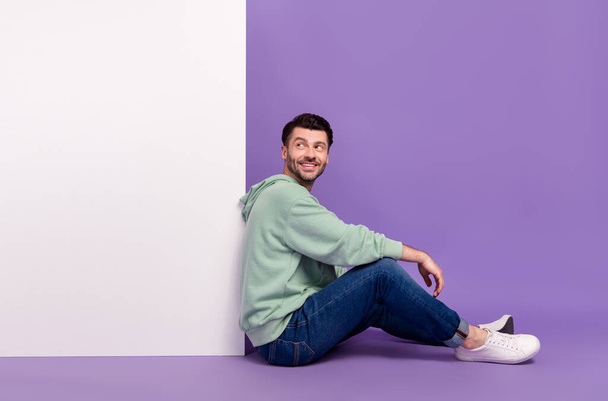 Täyspitkä kuva mukava nuori mies istua lattialla näyttää kiinnostunut valkoinen seinä pukeutunut tyylikäs khaki asu eristetty violetti väri tausta. - Valokuva, kuva