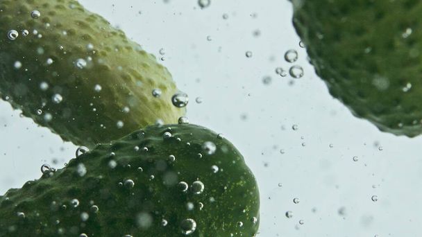 Closeup komkommers vallen vloeibaar in lichte achtergrond. Tuingroenten drijvend stijgende wateroppervlak. Groene biologische groenten wassen stroomt in glazen container. Ontspannende commerciële shot concept. - Foto, afbeelding