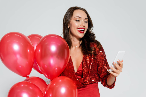 Cheerful and stylish woman holding balloons and using cellphone isolated on grey  - Φωτογραφία, εικόνα