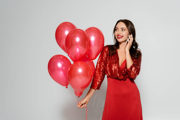 Веселая женщина в стильном красном платье разговаривает по мобильному телефону и держит воздушные шары на сером фоне  - Фото, изображение