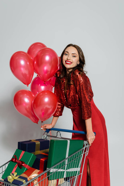 Happy and stylish woman standing near shopping cart with balloons and gifts on grey background  - Foto, immagini