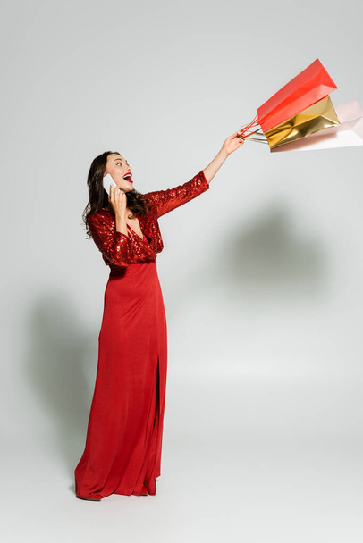 Excited woman in red dress talking on smartphone and holding shopping bags on grey background  - Foto, Imagen