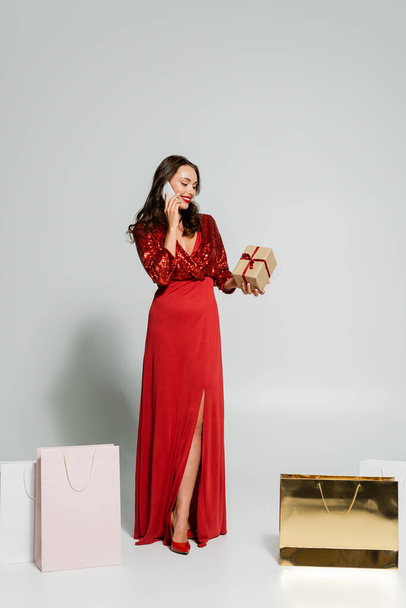 Pretty woman in red dress talking on smartphone and holding gift near shopping bags on grey background  - Foto, immagini