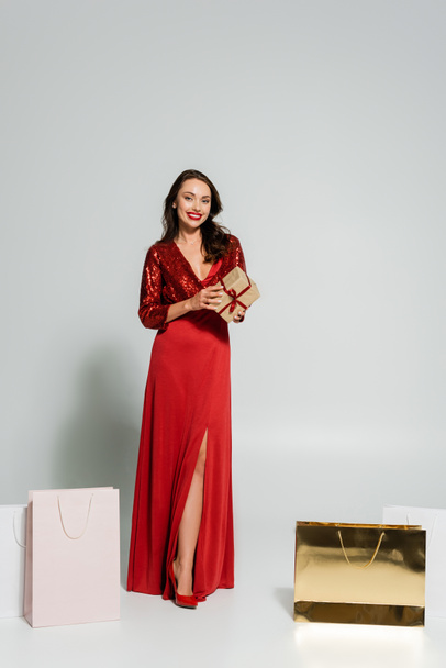 Lunghezza completa della donna alla moda in abito rosso contenente scatola regalo vicino a borse della spesa su sfondo grigio  - Foto, immagini