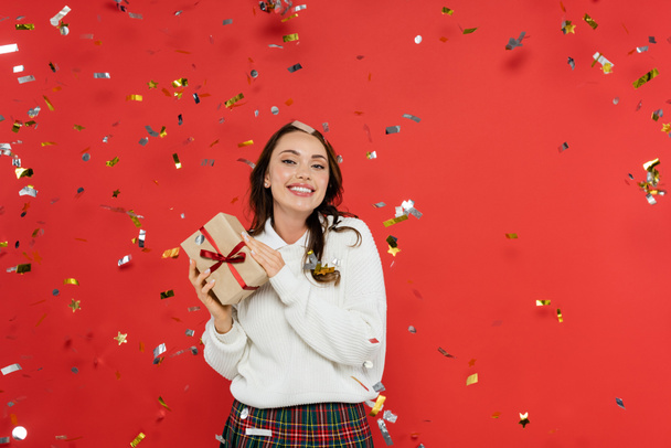 Cheerful brunette woman in sweater holding present under confetti on red background  - Fotó, kép