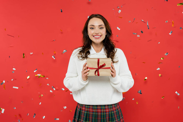 Cheerful brunette woman in warm sweater holding present with ribbon under confetti on red background  - Photo, Image