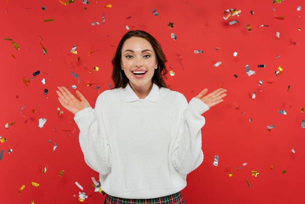 Excited brunette woman in warm sweater looking at camera under confetti on red background  - Фото, изображение