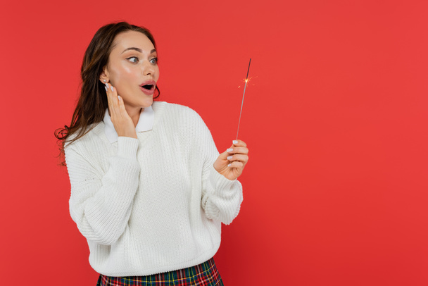 Shocked woman in sweater looking at sparkler isolated on red  - Photo, Image