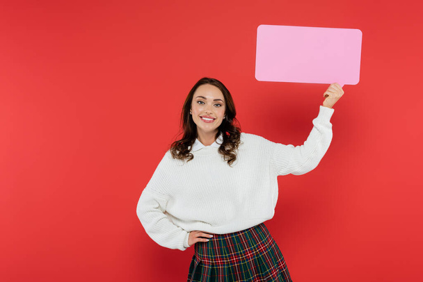 Positive brunette woman in sweater holding empty speech bubble on red background  - Photo, Image