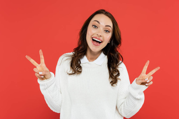 Positive brunette woman in white jumper showing peace sign isolated on red  - Foto, immagini