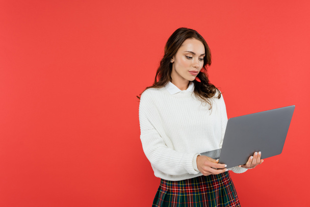 Brunette woman in warm jumper using laptop isolated on red  - Zdjęcie, obraz