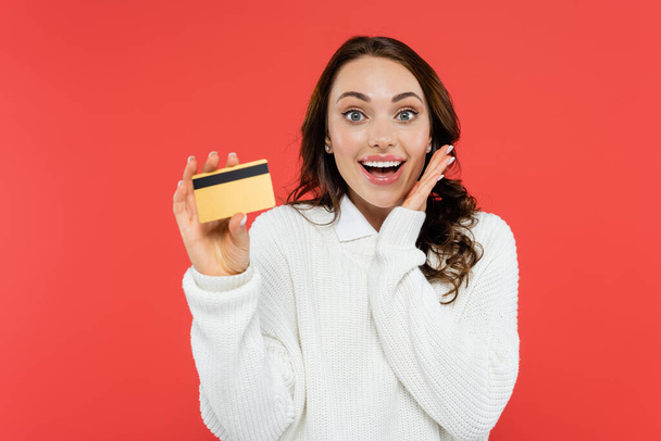 Excited brunette woman in white jumper holding credit card isolated on red  - Photo, image