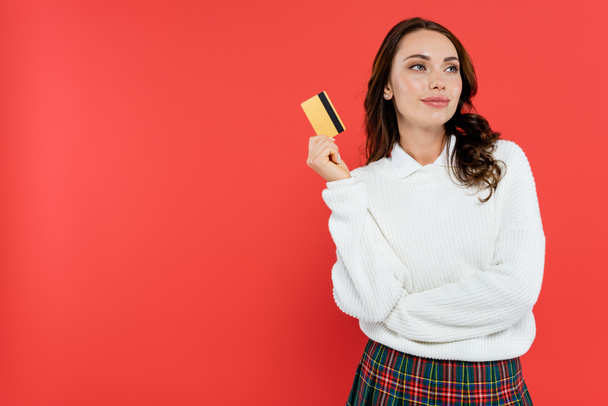 Dreamy brunette woman in cozy jumper holding credit card isolated on red  - Photo, Image