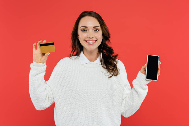 Pretty brunette woman in jumper holding cellphone and credit card isolated on red  - Fotografie, Obrázek