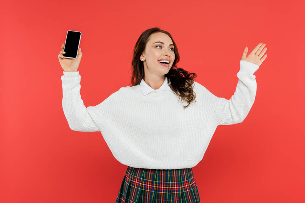 Cheerful young woman in cozy jumper holding smartphone isolated on red - Photo, Image