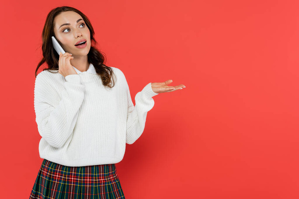 Brunette woman in warm sweater talking on mobile phone and pointing with hand on red background  - Foto, Imagem