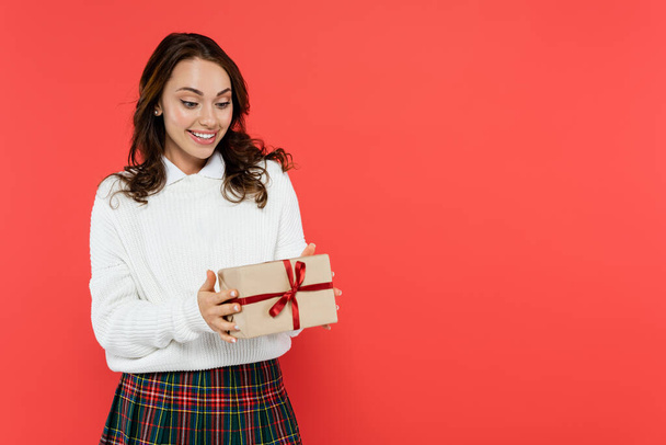 Positive young woman in warm sweater looking at gift isolated on red  - Φωτογραφία, εικόνα