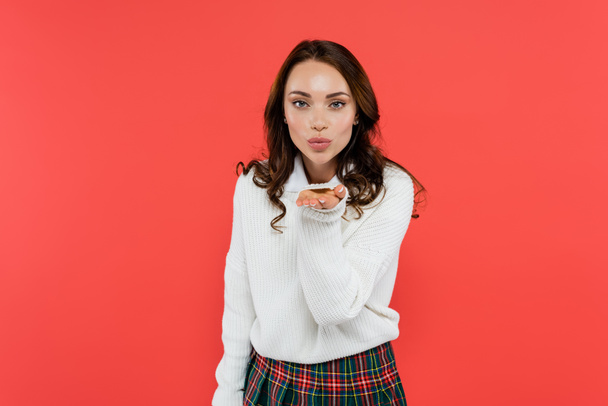 Brunette woman in jumper and plaid skirt blowing air kiss isolated on red  - Фото, зображення