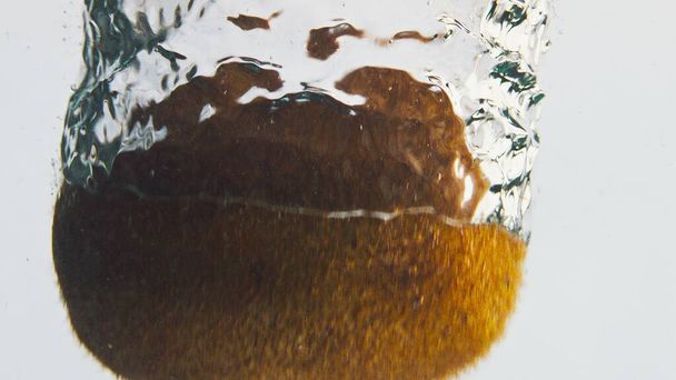 Gros plan kiwi chute d'eau en arrière-plan de lumière. Fruits tropicaux tombant faisant tourbillonner dans du liquide bouillonnant. Produit sucré à faible teneur en calories contenant en verre flottant. Belle publicité de délice de la cuisine. - Photo, image