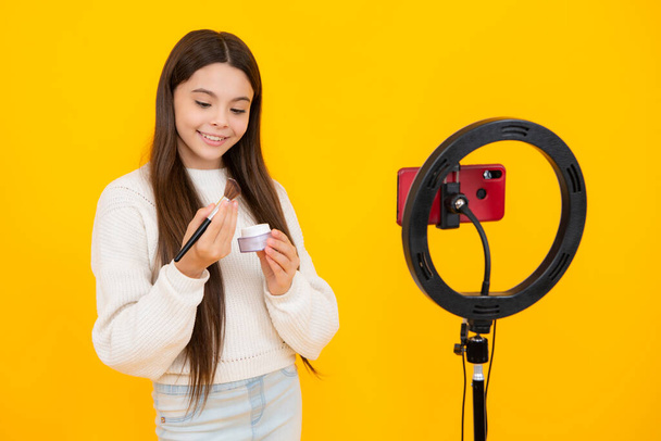 Beauty blog, presenting makeup cosmetics powder and brush. Teen girl speaking in front of camera for vlog isolated on yellow background. Teenager working as blogger, recording video blog - Photo, Image
