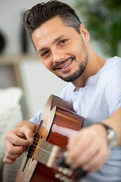 pohledný muž hraje na kytaru v obývacím pokoji - Fotografie, Obrázek