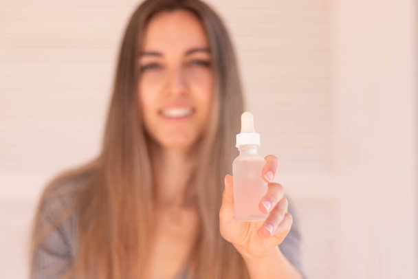 Вибірковий фокус білої і прозорої пляшки сироватки волосся в руці з розмитою молодою жінкою на білому тлі
 - Фото, зображення