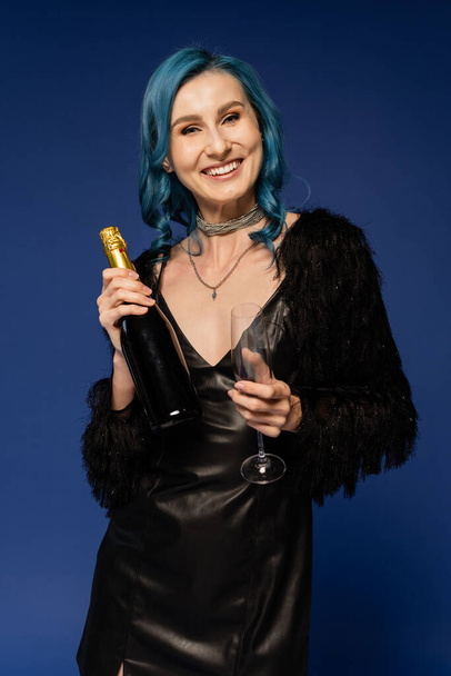mulher sorridente com cabelo colorido brilhante segurando vidro e garrafa de champanhe isolado em azul - Foto, Imagem