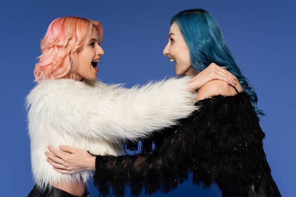 vista lateral de amigos queer sorprendidos en chaquetas de piel sintética en blanco y negro mirándose aislados en azul - Foto, imagen