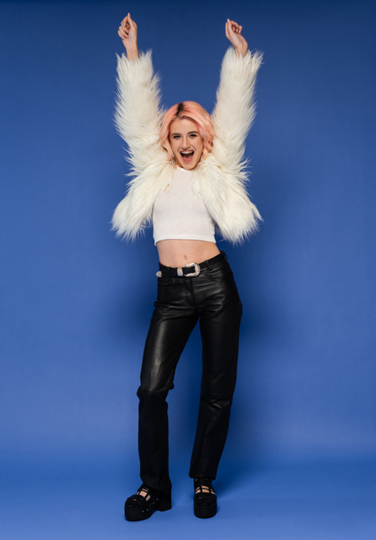 full length of excited woman in white faux fur jacket and black leather pants dancing with raised hands on blue background - Photo, Image