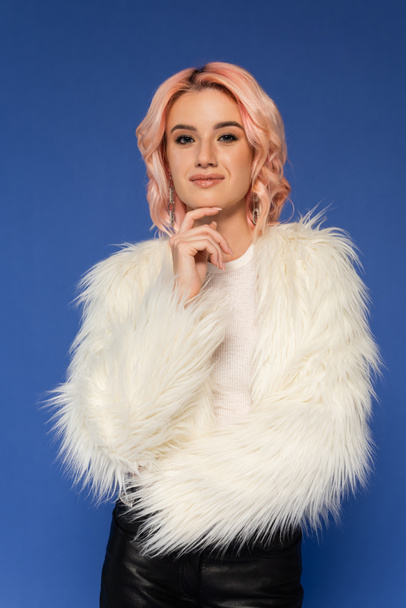 positive and dreamy woman in white faux fur jacket holding hand near face and looking at camera isolated on blue - Foto, afbeelding