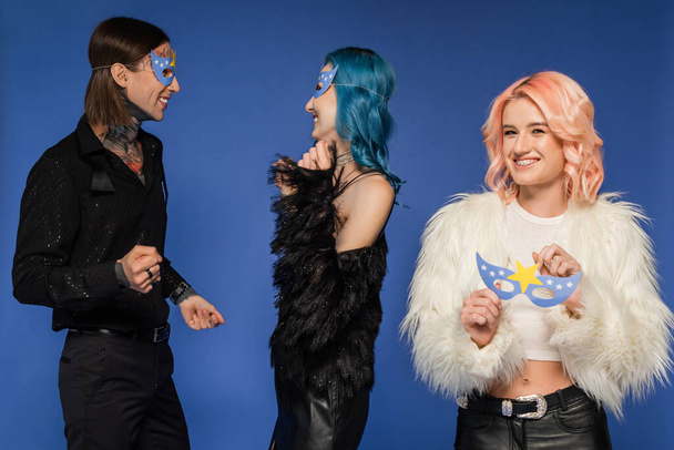 queer persoon holding party masker en kijken naar camera in de buurt van niet-binaire mensen dansen geïsoleerd op blauw - Foto, afbeelding