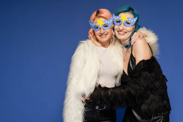 alegre queer personas en partido máscaras y chaquetas de piel sintética abrazando aislado en azul - Foto, Imagen
