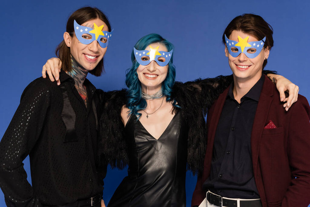 feliz persona queer en vestido de cuero negro abrazando amigos no binarios en máscaras de fiesta aislados en azul - Foto, imagen
