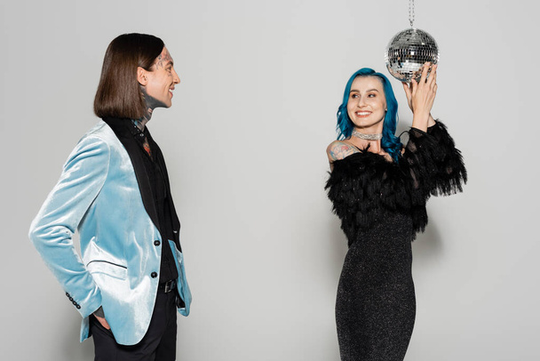 uśmiechnięta queer osoba w niebieskiej peruce dotykając disco ball obok elegancki nonbinary przyjaciel na szarym tle - Zdjęcie, obraz
