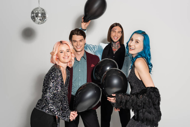 pleased queer people with black festive balloons smiling at camera on grey background - Zdjęcie, obraz