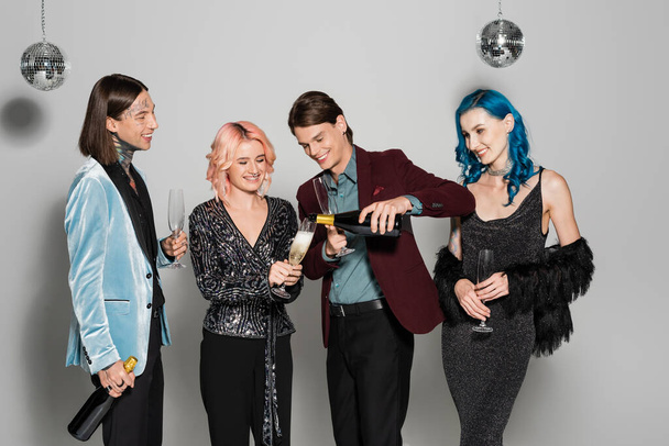 souriant queer personne versant du champagne pendant la fête de Noël avec des amis élégants non binaires sur fond gris - Photo, image