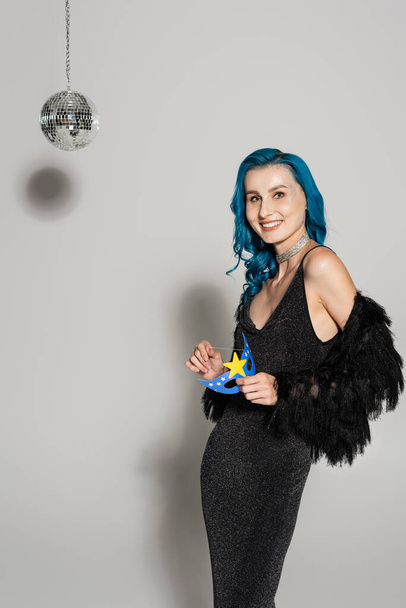 elegant woman with blue hair holding party mask and smiling at camera near disco ball on grey background - Foto, Bild