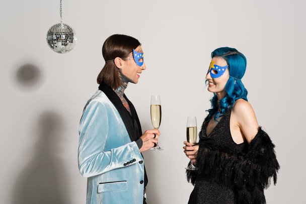 Seitenansicht stilvoller queerer Freunde mit Champagnergläsern in der Nähe der Discokugel auf grauem Hintergrund - Foto, Bild