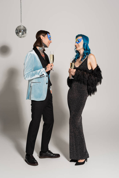 longitud completa de elegantes amigos queer en máscaras de fiesta con copas de champán sobre fondo gris - Foto, Imagen