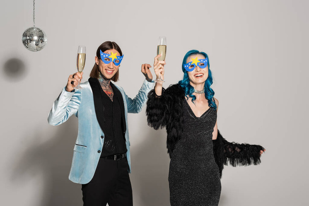 stylish nonbinary friends in party masks toasting with champagne while smiling at camera on grey background - Photo, Image