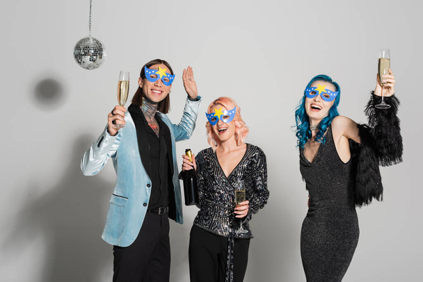 tattooed nonbinary person holding champagne glass and waving hand near nonbinary friends in party masks on grey background - Photo, Image