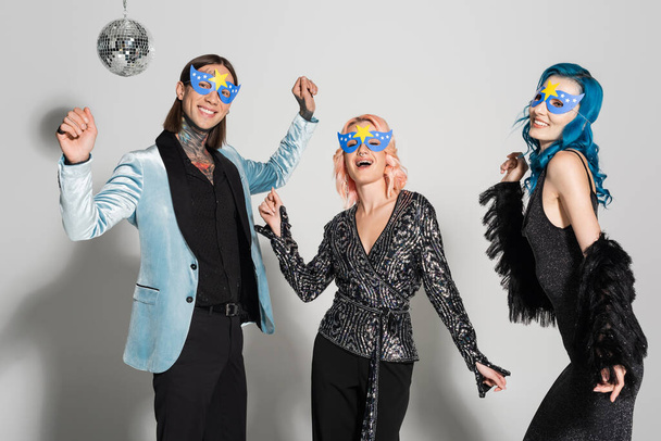 happy queer people in elegant clothes and party masks dancing during new year party on grey background - Фото, зображення
