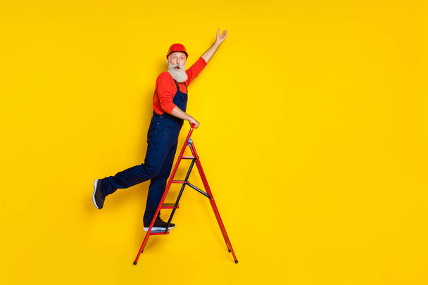 Full length photo of positive funky age man wear overall uniform red hard hat walking ladder empty space isolated yellow color background. - Photo, image