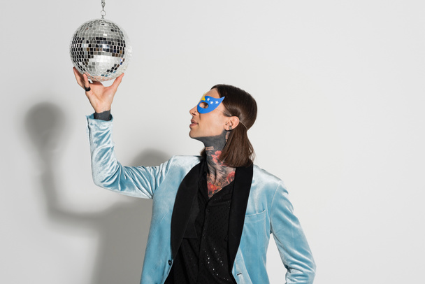 queer person in elegant silk blazer and party mask touching shiny disco ball on grey background - Foto, imagen